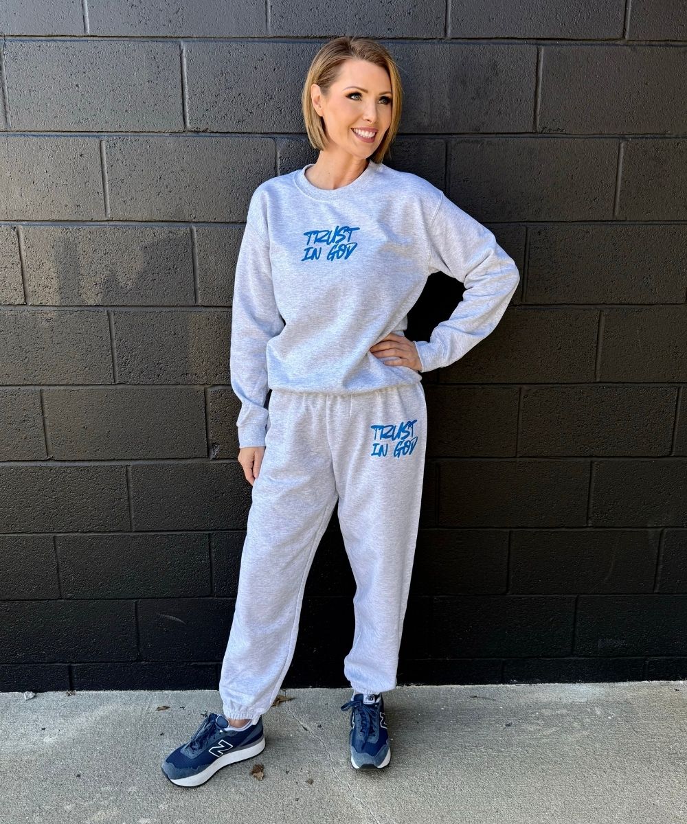 Person wearing a white sweatshirt and pants with text, standing against a black brick wall.