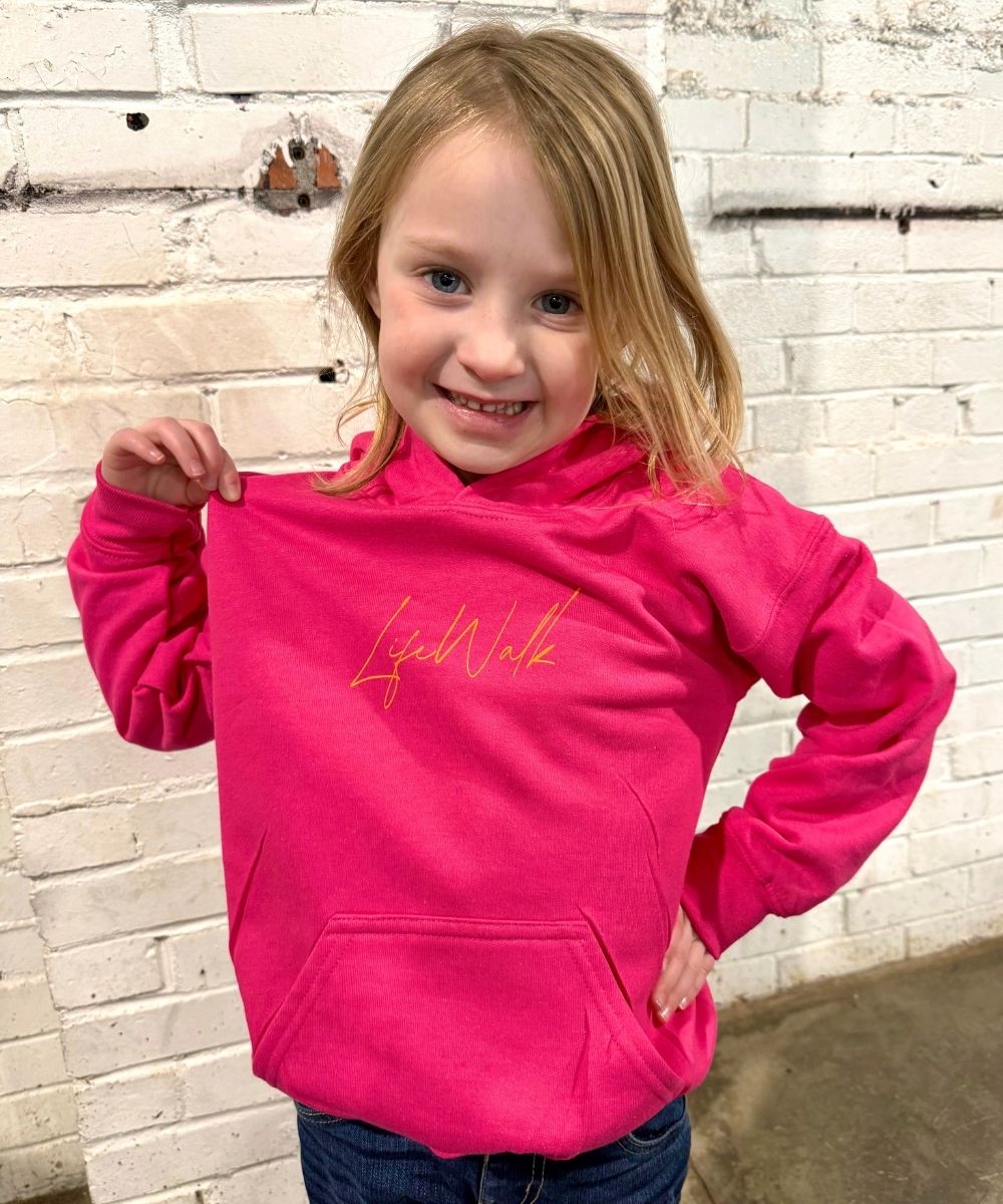 Child wearing a bright pink hoodie with text, standing against a light brick wall.