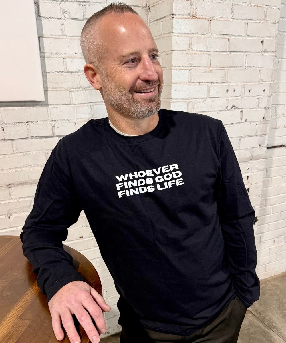 Man wearing a black long-sleeve shirt with text, sitting against a white brick wall.