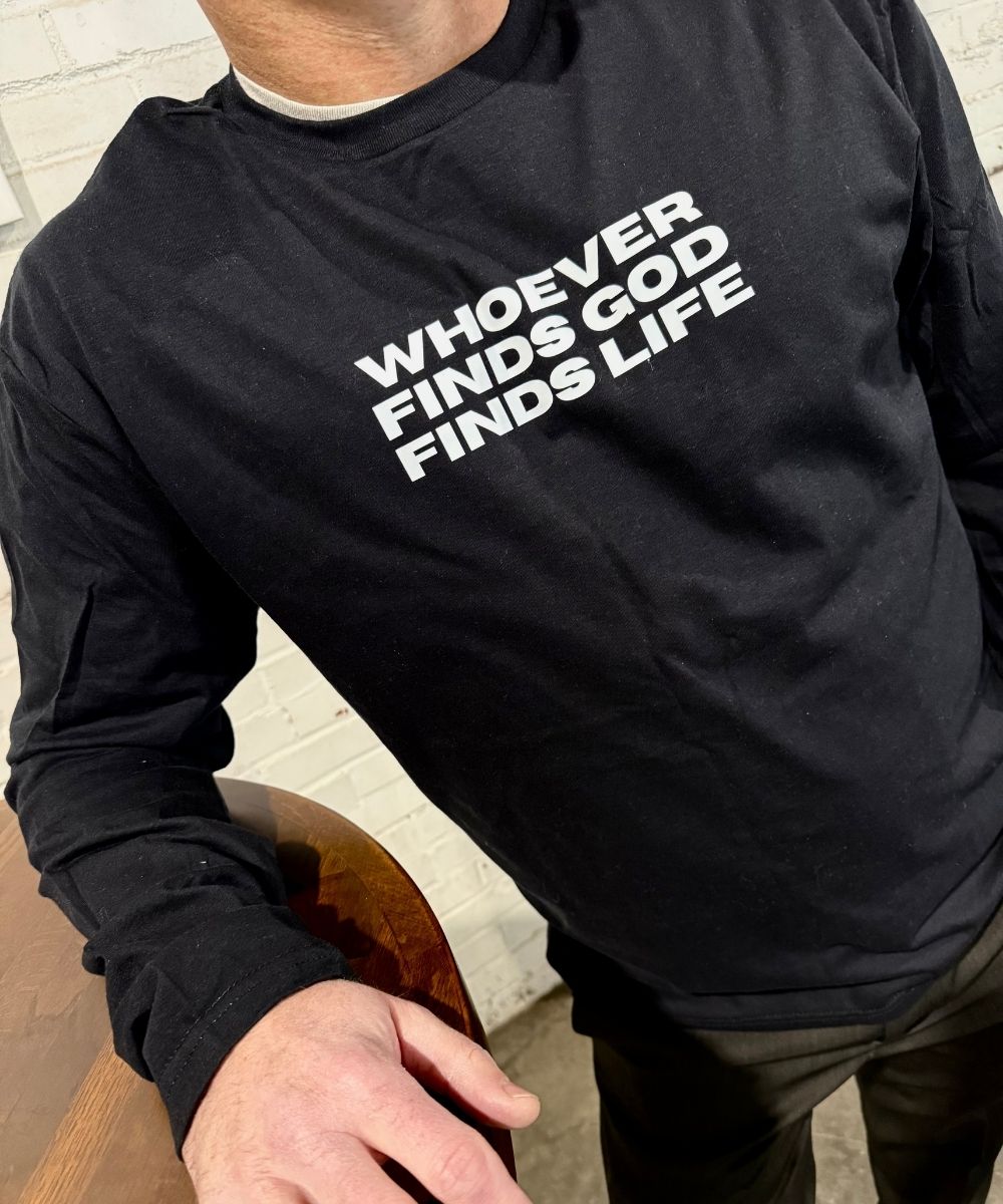 Person wearing a black sweatshirt with text 'WHOEVER GOD FINDS GOD FINDS LIFE' in a casual setting.