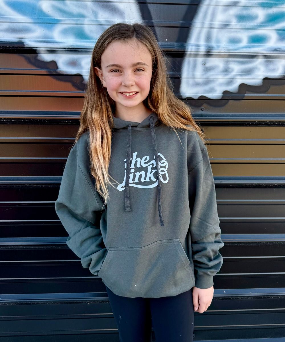 Young girl wearing a gray hoodie with a logo, standing in front of a graffiti-covered wall.