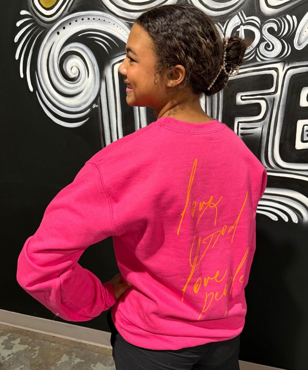 Person wearing a bright pink sweatshirt with text on the back, standing against a decorative wall.