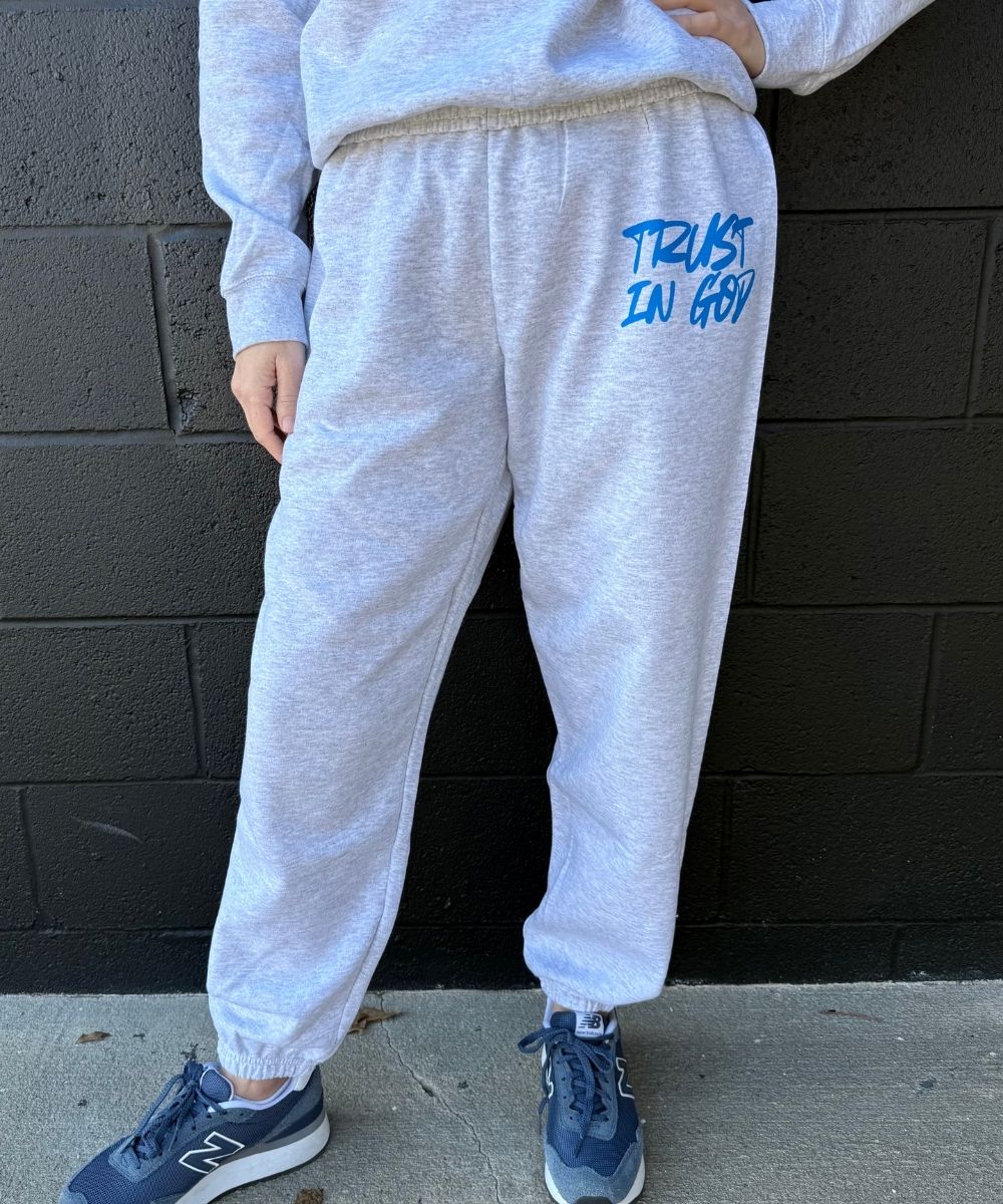 Person wearing gray sweatpants with 'Trust In God' text, standing against a black brick wall.